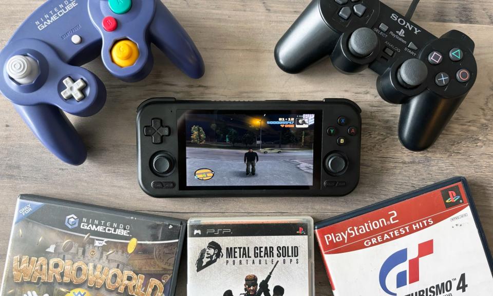 A black handheld gaming device, the Retroid Pocket 4 Pro, sits on a brown wooden table surrounded by various video games and gamepads, including controllers for the Sony PlayStation 2 and Nintendo GameCube.