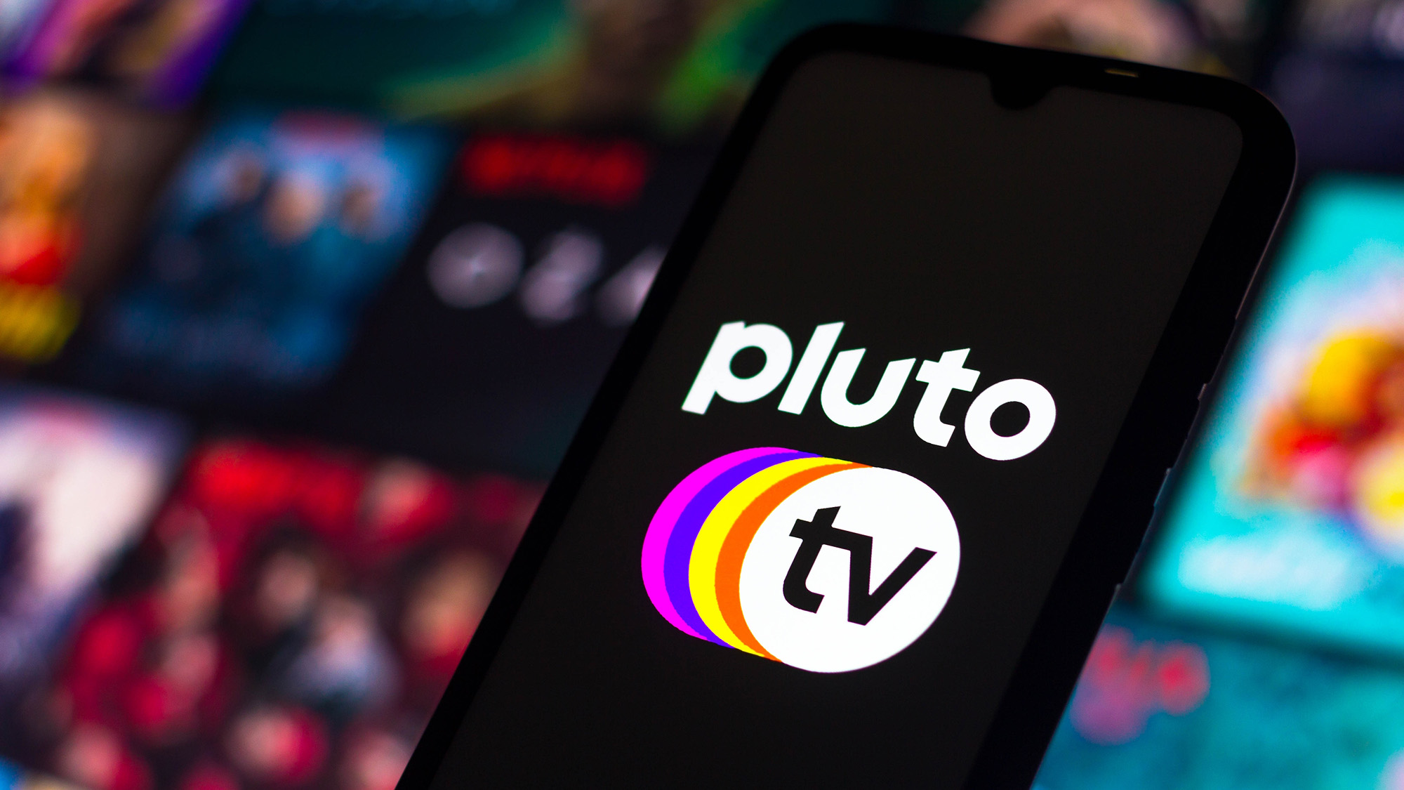 Το λογότυπο Pluto TV φαίνεται σε ένα τηλέφωνο μπροστά από μια οθόνη θολών εικόνων τηλεοπτικών εκπομπών